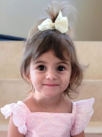 little girl with brown hair and brown eyes wearing a pink ruffle shirt and white bow in her hair