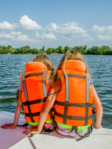 The backs of two sisters wearing life vests sitting on the dock of the lake